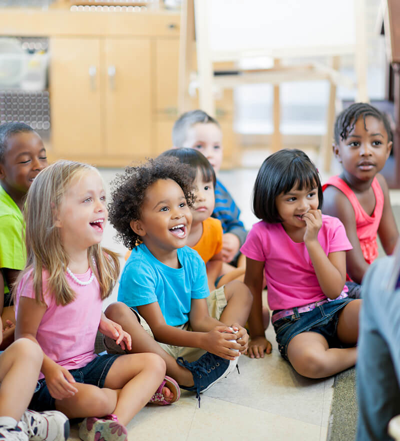 A group of children sitting on the floor enjoying listening to a teacher.